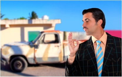 A smarmy car salesman with a junk pickup behind him.