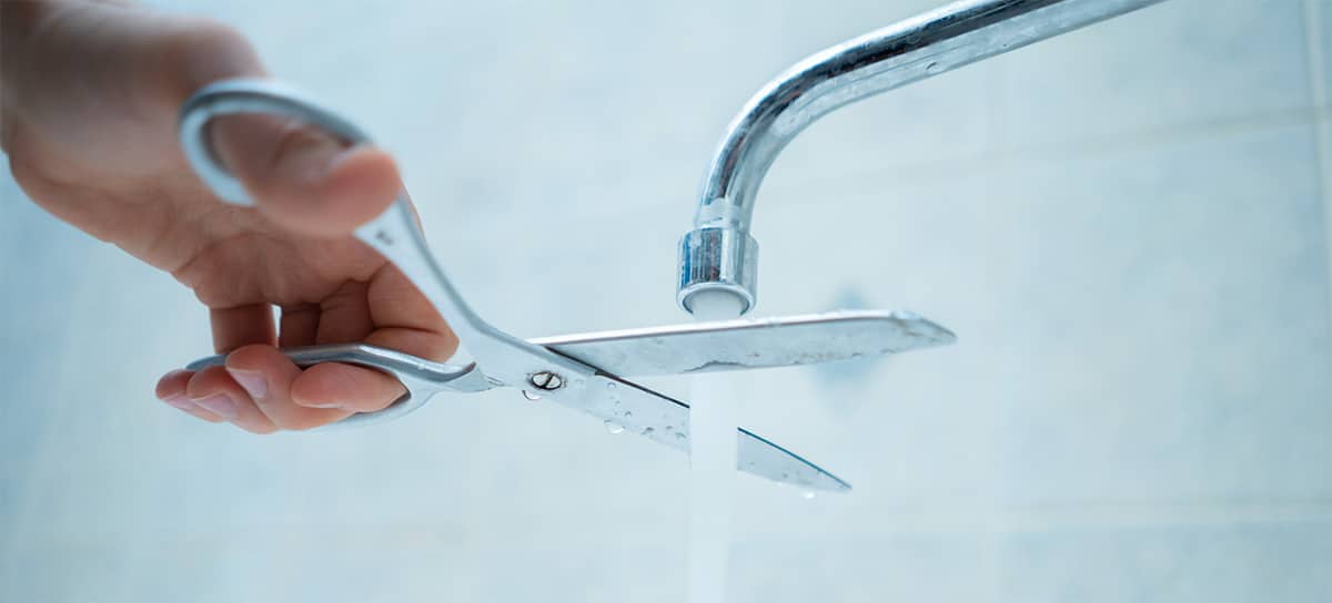 A hand with a pair of scissors snapping at a running faucet's water.
